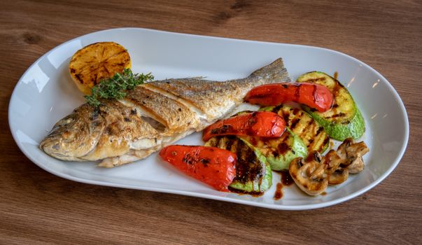 Plat de poisson grillé accompagné de légumes colorés, courgettes, poivrons et champignons sur un fond en bois.